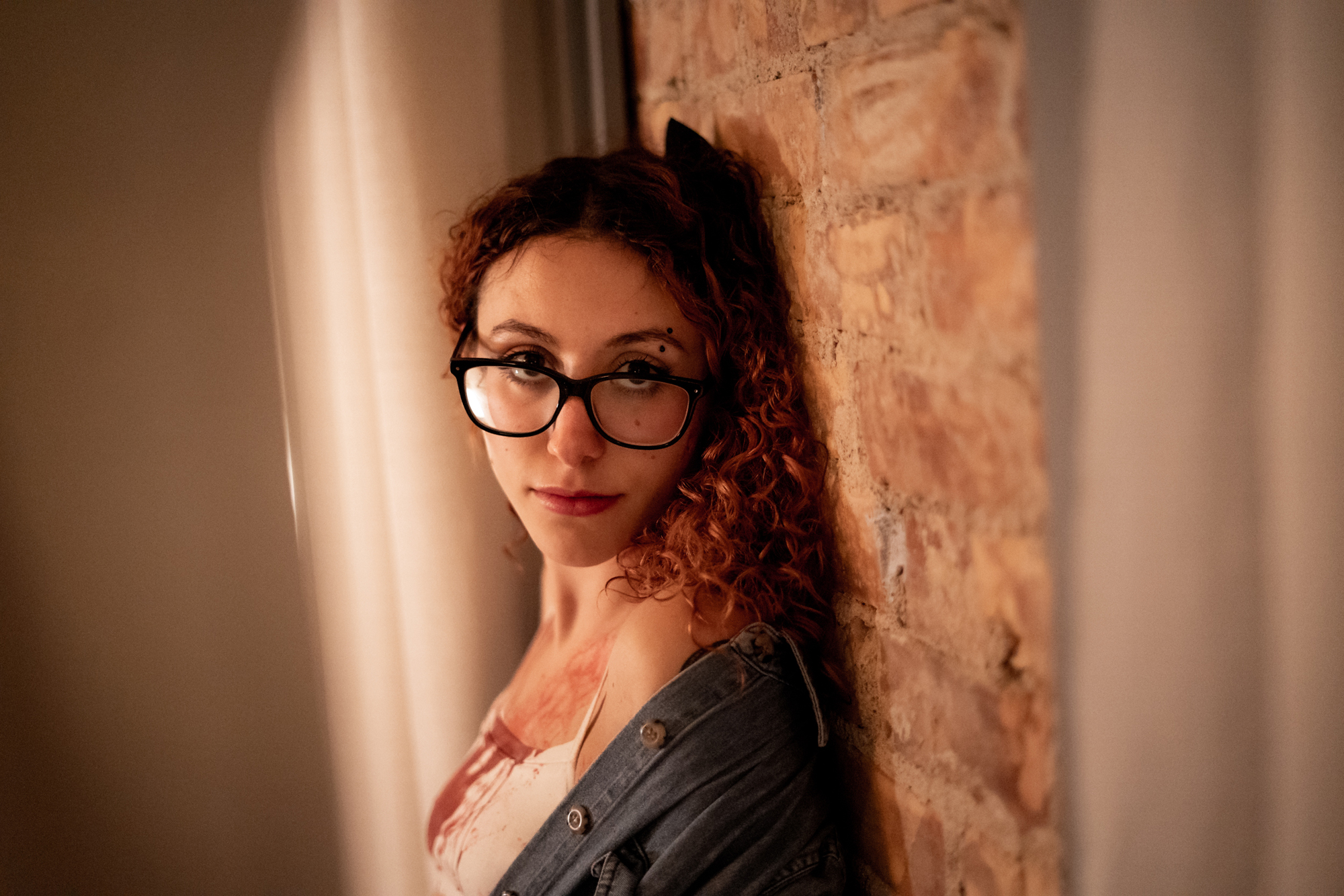 Best boudoir drama: Beautiful woman in white bra and panty lingerie, adorned with a jean jacket, leaning against a dripping (faux) blood-stained brick wall, framed by a curtain for an alluring and provocative scene.