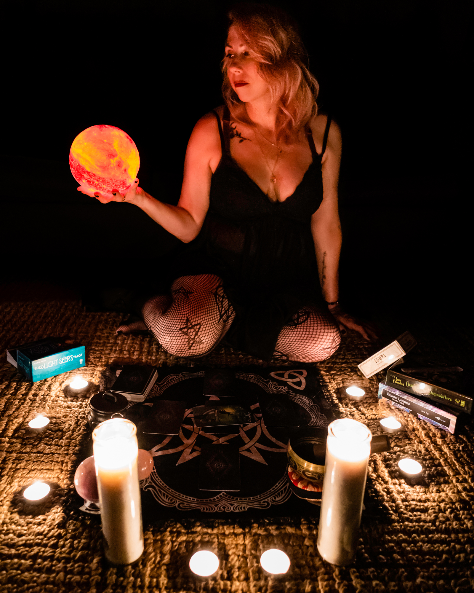 Best boudoir mystique: Woman in lingerie using a cloth altar for tarot, surrounded by candles, holding an illuminating orange orb in a darkened room.